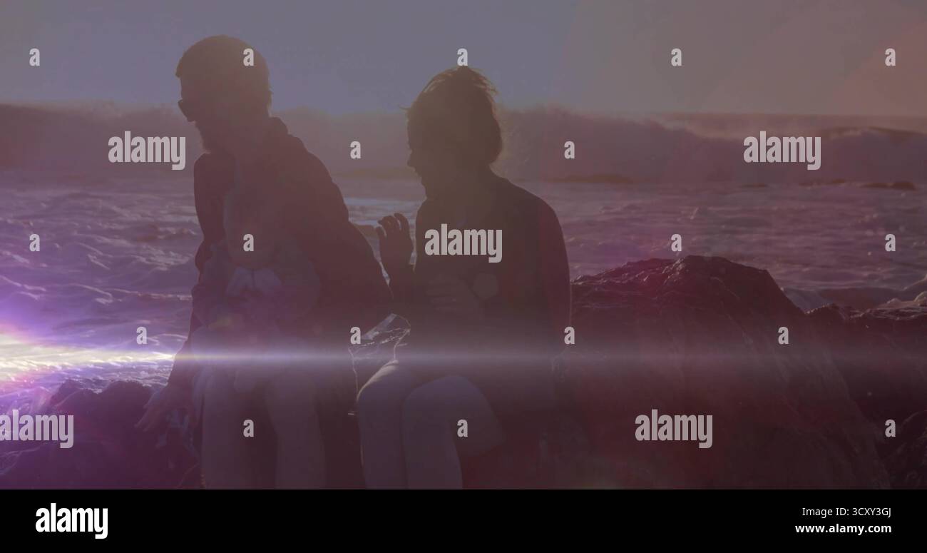 Homme assis portant veste et casquette, femme tenant bébé sur la roche du rivage au coucher du soleil, vagues de l'océan Banque D'Images