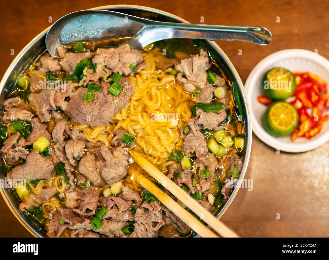 Cuisine traditionnelle du Vietnam, servie dans un bol en métal et une cuillère à soupe, avec des oignons verts frais, de l'ail et des nouilles de riz jaune dans un bouillon épicé, w Banque D'Images