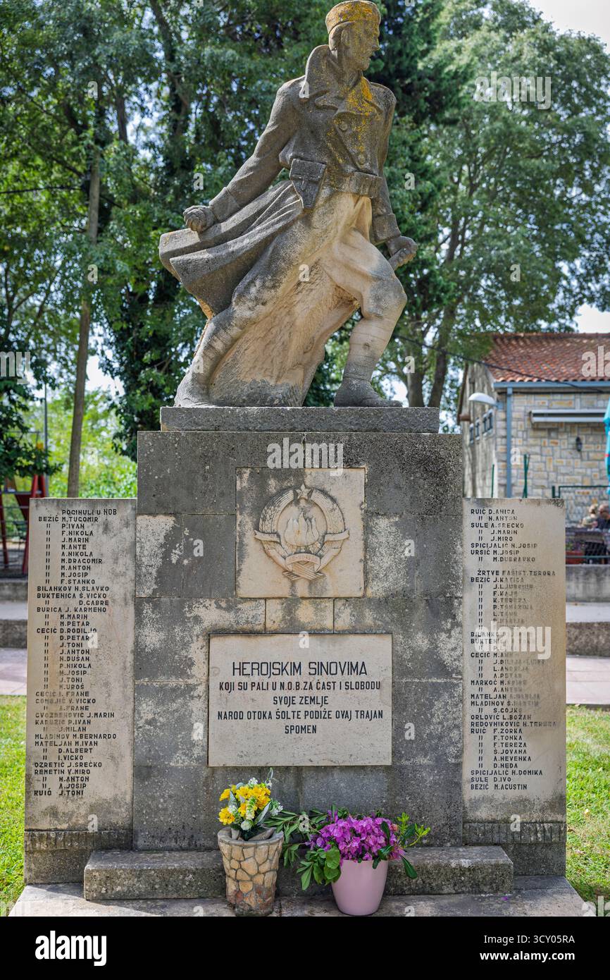 Grohote, CROATIE, 08.29.2025 :Statue en pierre extérieure d'un soldat en marche au sommet d'un piédestal de brique, flanqué de plaques nominatives. Des fleurs fraîches reposent à la base Banque D'Images