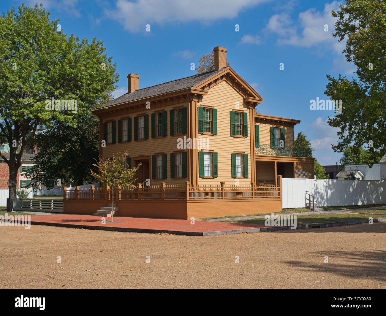 Maison historique à ossature jaune avec volets verts et porche enveloppant se trouve au milieu des arbres et des clôtures blanches sous un ciel partiellement nuageux. Ce puits... Banque D'Images