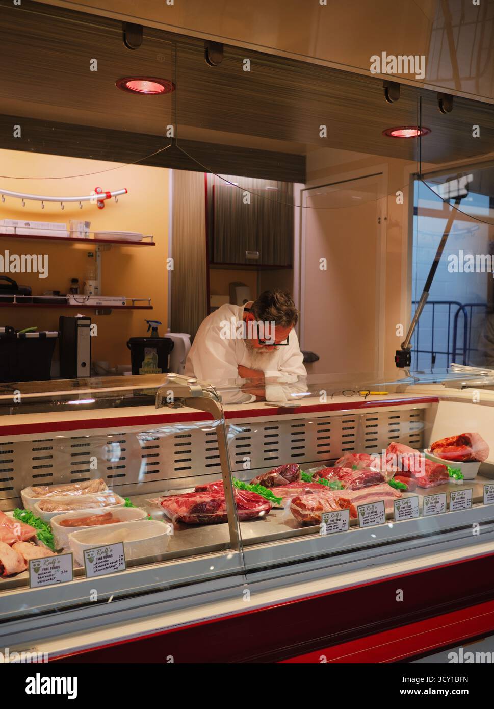 Un boucher masculin vêtu d'un manteau blanc repose derrière son comptoir de fines coupes ; la lumière naturelle et tamisée suggère l'habileté et une petite entreprise de confiance. Banque D'Images