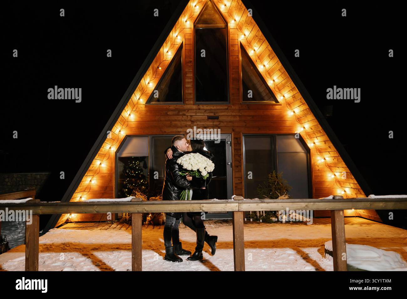 Couple s’embrassant avec un bouquet de roses blanches devant un chalet en bois décoré de lumières lors d’une nuit d’hiver Banque D'Images