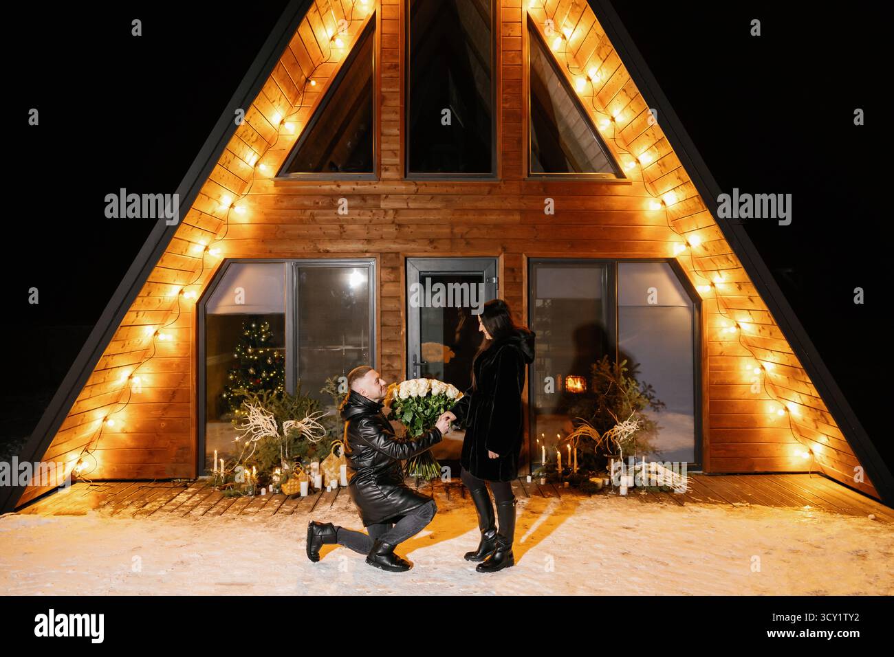 Surprise romantique avec des roses blanches devant une maison en bois confortable et illuminée en hiver Banque D'Images