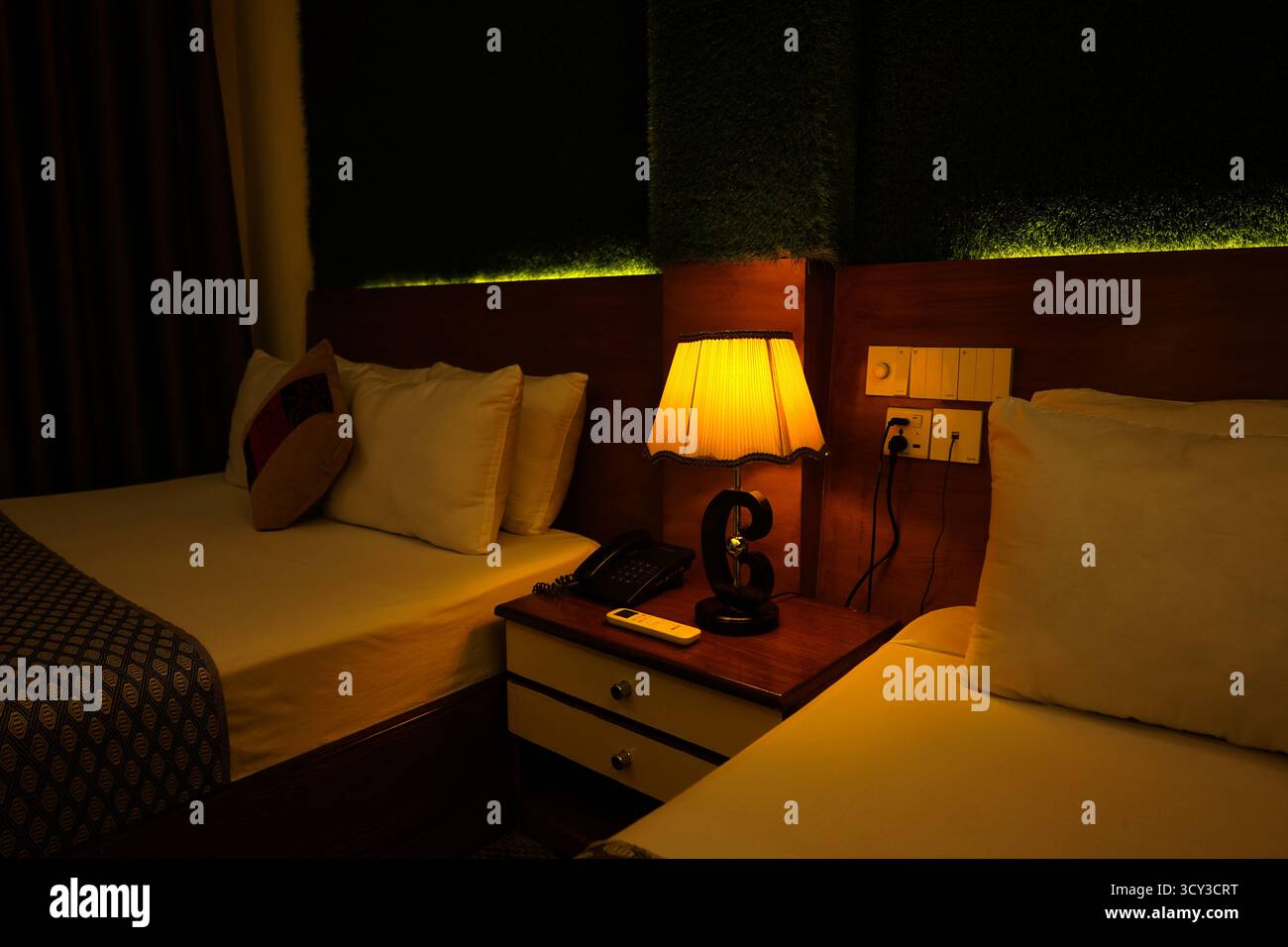 Une image de faible luminosité d'une table de nuit de chambre d'hôtel entre deux lits, avec une lampe de table jaune lumineux, un téléphone et une télécommande, créant un AT confortable Banque D'Images