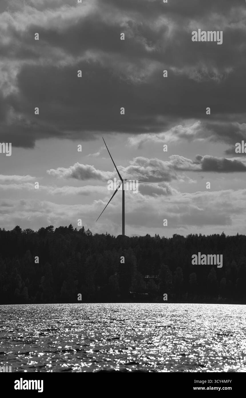 Une éolienne se lève derrière Une forêt verte sur la rive du lac. La lumière du soleil se reflète sur l'eau, les nuages dérivent à travers le ciel. Un symbole de Sustainable en Banque D'Images