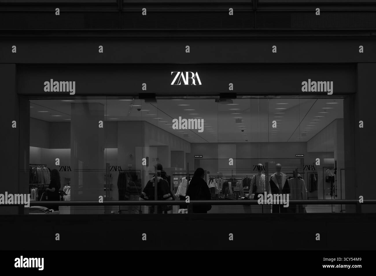 Image en noir et blanc de clients marchant devant un magasin Zara, capturant la vie moderne du commerce de détail et la mode de rue dans une composition minimaliste. Banque D'Images