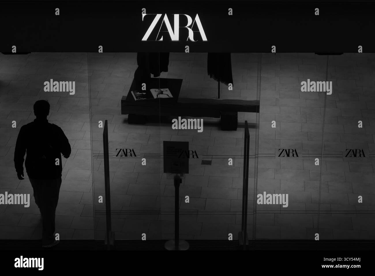 Image en noir et blanc d'un homme entrant dans un magasin Zara, capturant la vie moderne du commerce de détail et la mode de rue dans une composition minimaliste. Banque D'Images