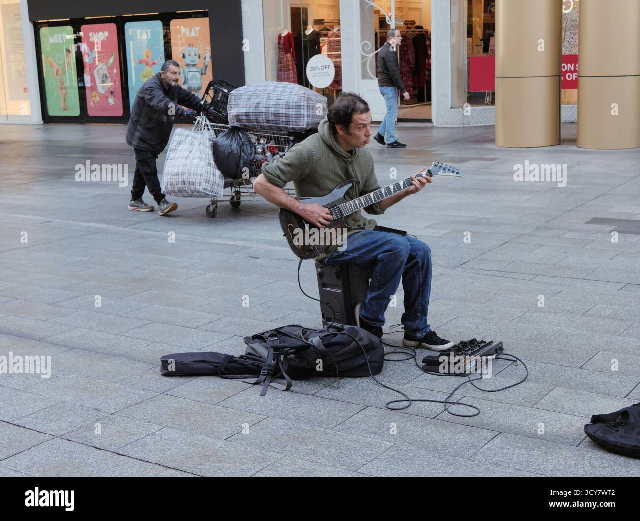 Guitariste solo assis sur un ampli, jouant de la musique dans un quartier commerçant ; moment réaliste, franc, véhiculant un sentiment de talent et de grit. Banque D'Images