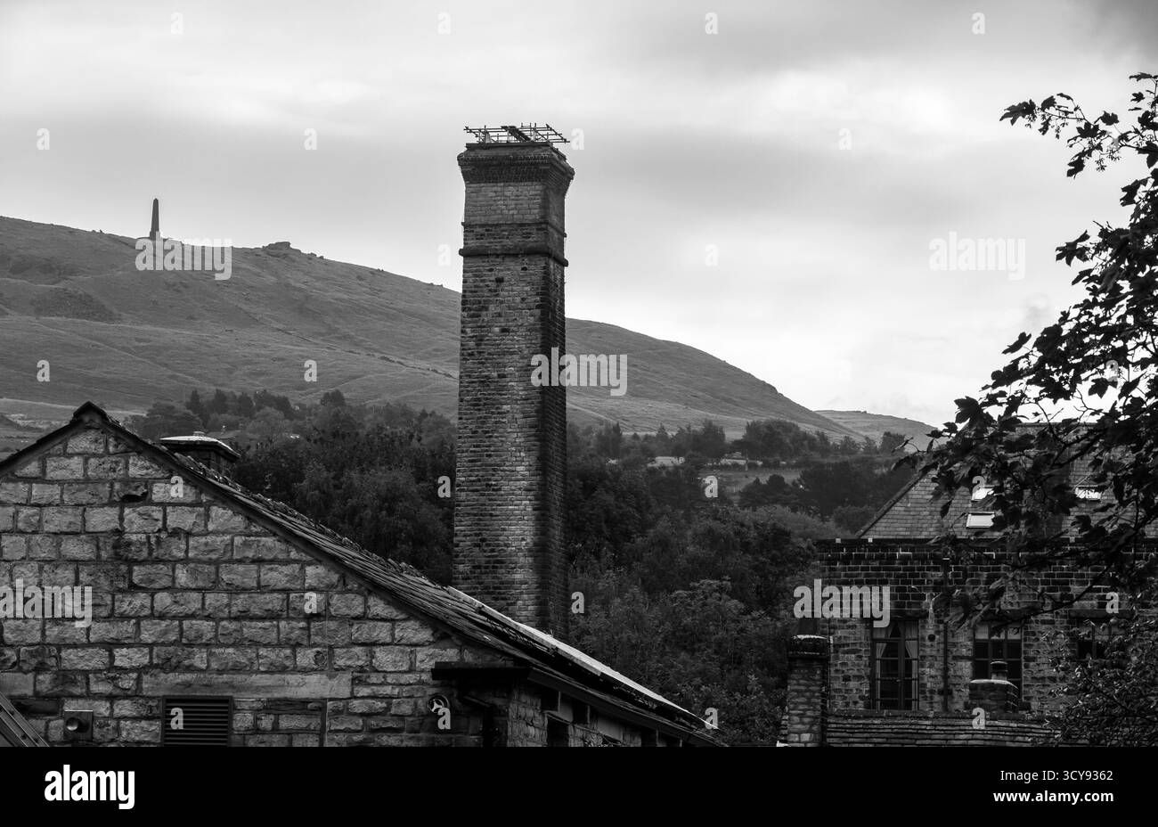 Des bâtiments historiques de moulin en pierre et une grande cheminée avec des collines en arrière-plan, y compris le monument commémoratif de guerre pots and pans au-dessus de Uppermill en monotone Banque D'Images