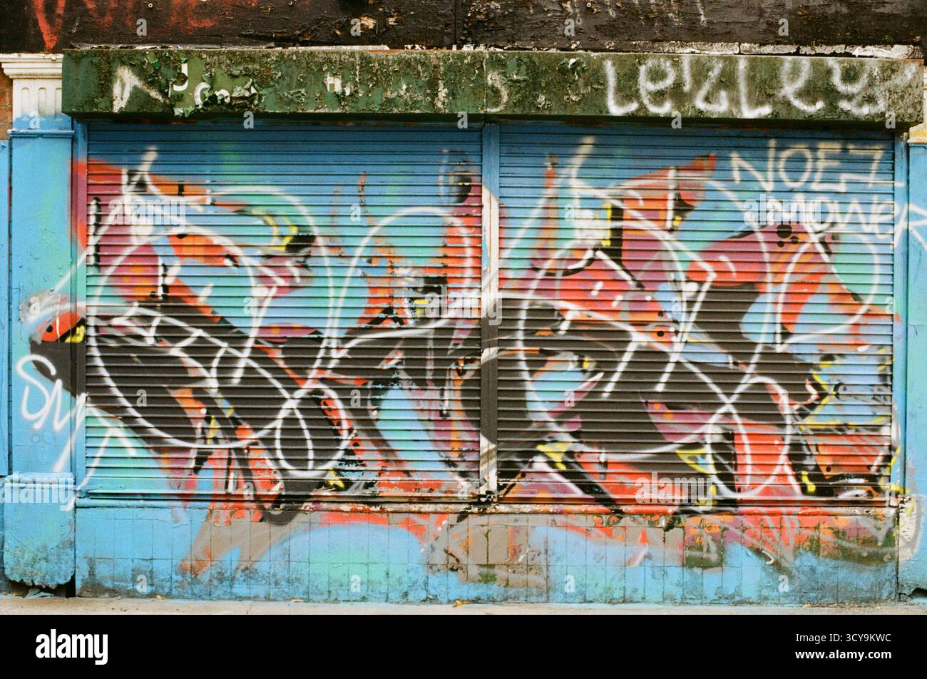 Graffiti coloré aux tons bleu, orange et rose sur un obturateur de magasin à Manchester, en Angleterre, photographié sur film couleur Fujifilm 200 Banque D'Images