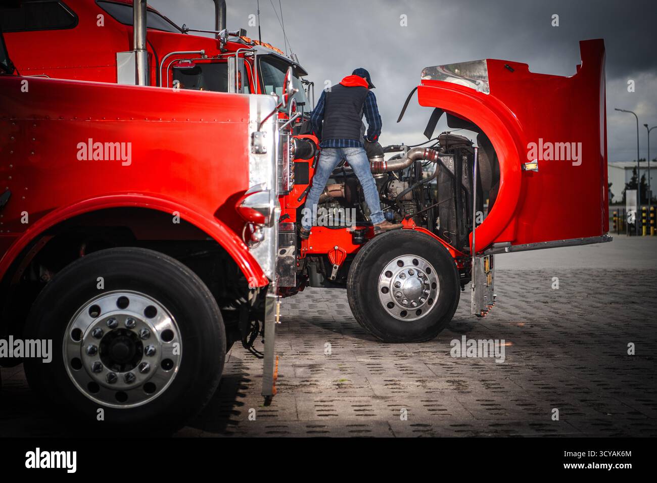 Un mécanicien travaille sur le moteur d'un camion rouge dans un centre de transport sous ciel nuageux pendant l'après-midi. Banque D'Images