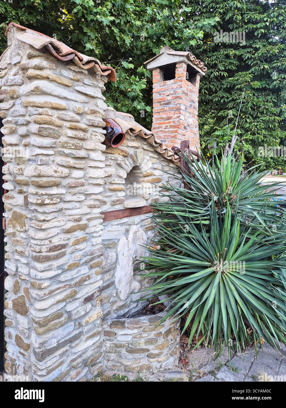 un poêle extérieur en pierre à côté d'un vaste buisson de yucca à l'ombre des arbres du jardin. Confort méditerranéen - Image de stock capturée avec un smartphone