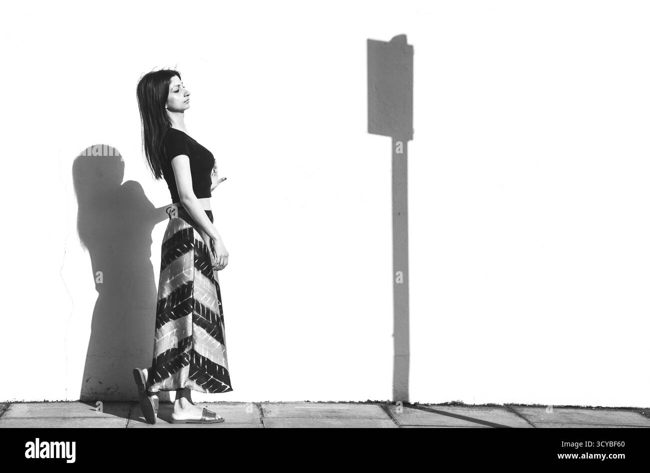 Femme debout à la lumière du soleil projetant une longue ombre sur un mur blanc. Banque D'Images