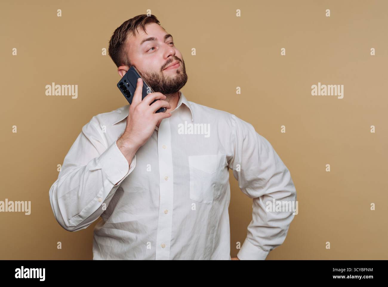 Un homme portant une chemise blanche se tient debout avec une main sur sa hanche, tenant un smartphone à son oreille. Il semble réfléchi et engagé dans la conversation, Creat Banque D'Images