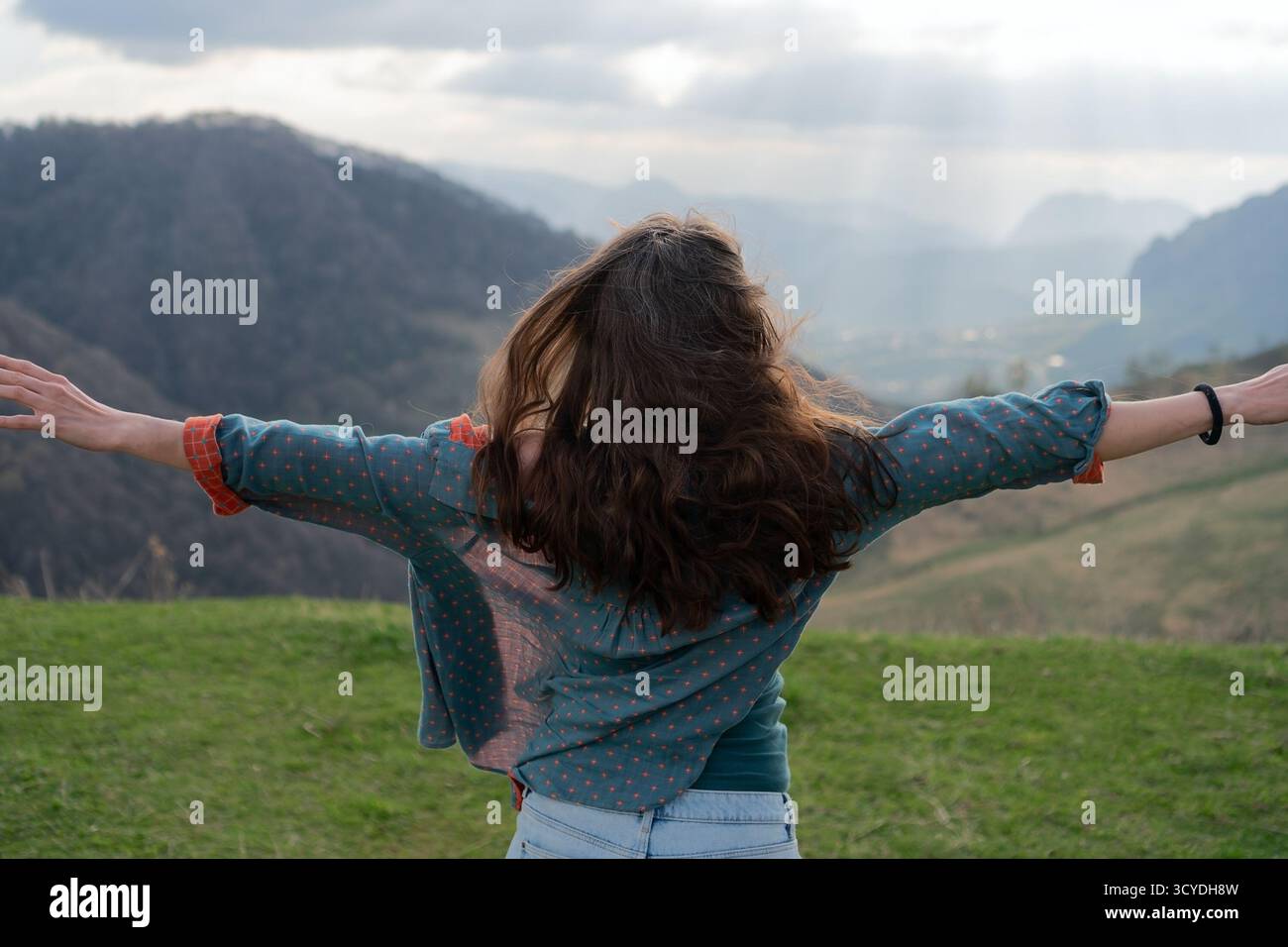 Vue arrière de la jeune femme en short et chemise debout au bord de la montagne, les bras tendus dans des conditions venteuses. Le temps nuageux rehausse les paysages spectaculaires. Banque D'Images