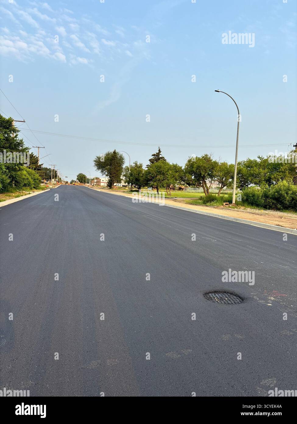 Rue résidentielle fraîchement pavée, montrant une nouvelle surface lisse en asphalte, des arbres en bordure de route et des lignes électriques dans un quartier de banlieue. - Image de stock capturée avec un smartphone