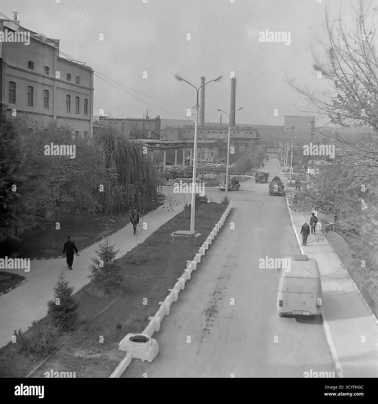 Cette photo d'archives en noir et blanc des années 1970 donne une vue verticale et en angle élevé de l'armature-isolateur Slavyansk (AIZ) à Sloviansk, URSS. La photo donne sur une longue route d'usine et une allée piétonne parallèle bordée d'arbres. Les travailleurs marchent sur les chemins, et les véhicules, y compris une camionnette (type UAZ/RAF) et des camions, sont sur la route. En arrière-plan, le paysage industriel présente un vieil atelier de briques et deux cheminées hautes. Un panneau publicitaire de propagande est également visible Banque D'Images