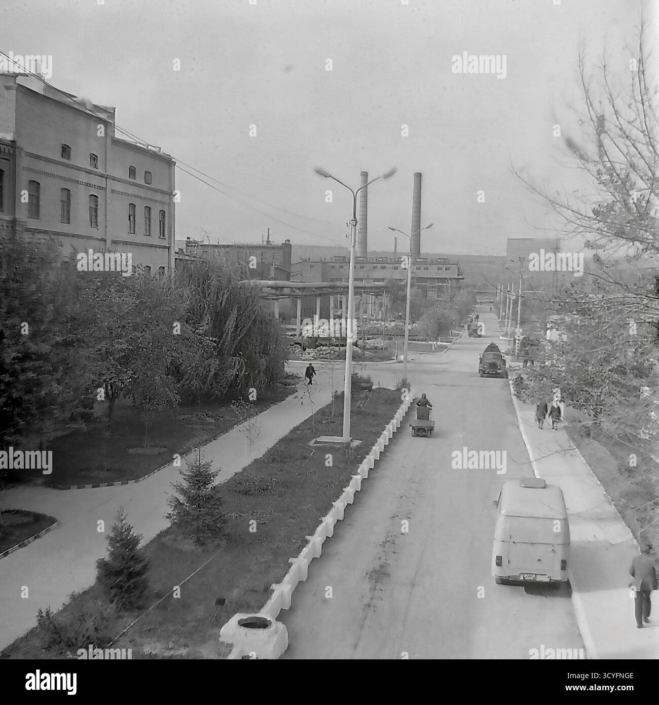 Cette photo d'archives en noir et blanc des années 1970 donne une vue verticale et en angle élevé de l'armature-isolateur Slavyansk (AIZ) à Sloviansk, URSS. La photo donne sur une longue route d'usine et une allée piétonne parallèle bordée d'arbres. Les travailleurs marchent sur les chemins, et les véhicules, y compris une camionnette (type UAZ/RAF) et des camions, sont sur la route. En arrière-plan, le paysage industriel présente un vieil atelier de briques et deux cheminées hautes. Un panneau publicitaire de propagande est également visible Banque D'Images