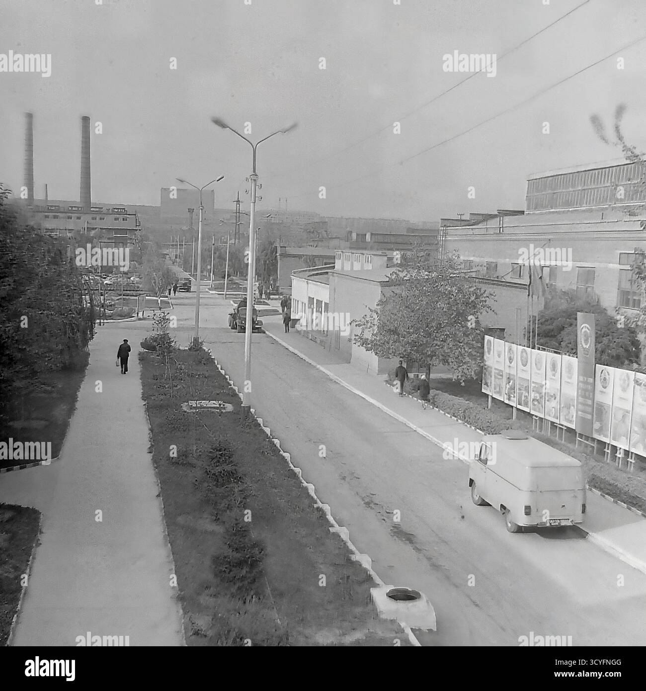 Cette photo d'archives en noir et blanc des années 1970 donne une vue verticale et en angle élevé de l'armature-isolateur Slavyansk (AIZ) à Sloviansk, URSS. La photo donne sur une longue route d'usine et une allée piétonne parallèle bordée d'arbres. Les travailleurs marchent sur les chemins, et les véhicules, y compris une camionnette (type UAZ/RAF) et des camions, sont sur la route. En arrière-plan, le paysage industriel présente un vieil atelier de briques et deux cheminées hautes. Un panneau publicitaire de propagande est également visible Banque D'Images