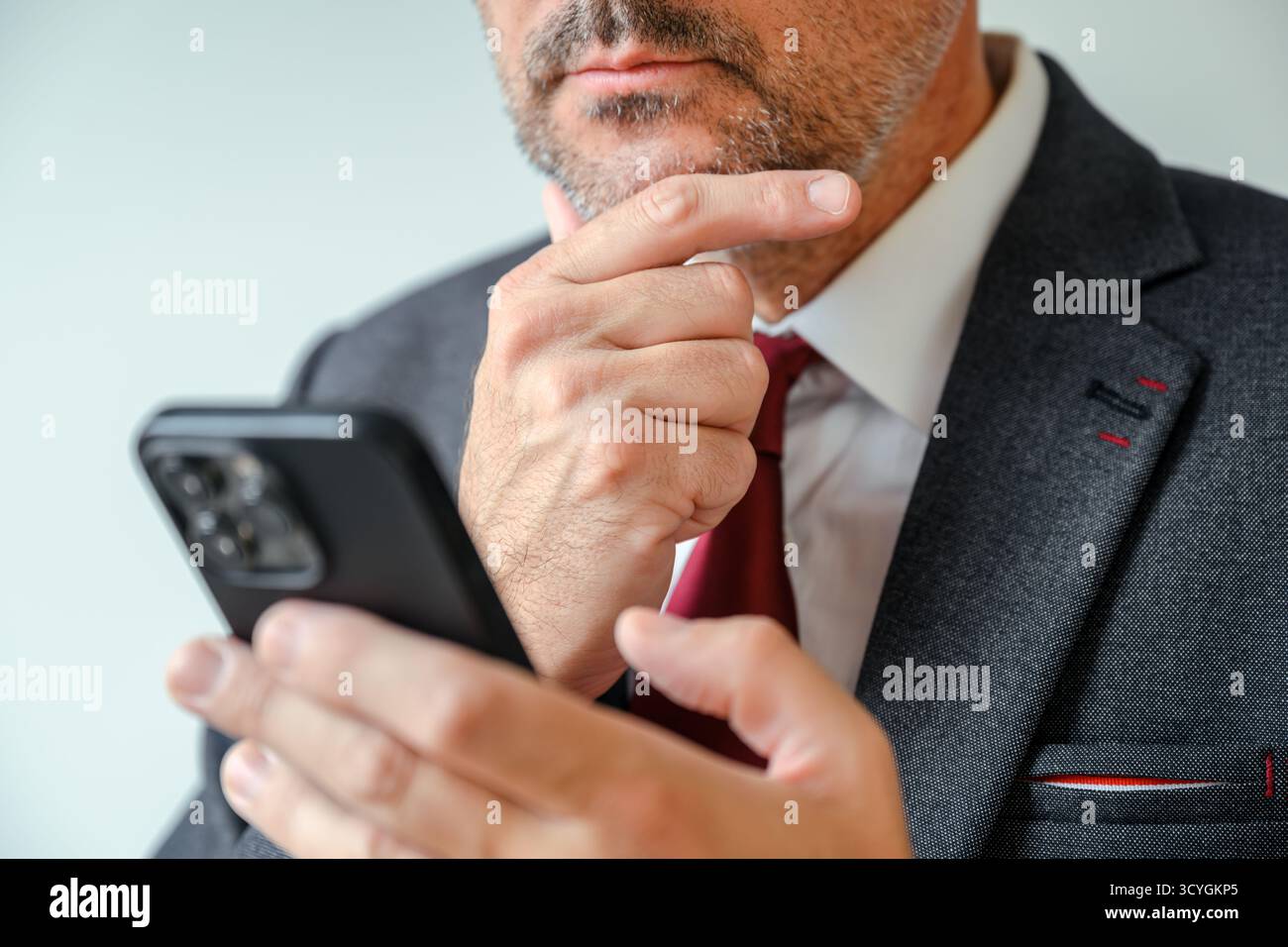 Homme d'affaires contemplatif tenant un smartphone et lisant un message texte avec la main sur le menton, moment de bureau réfléchi. Mise au point sélective. Banque D'Images