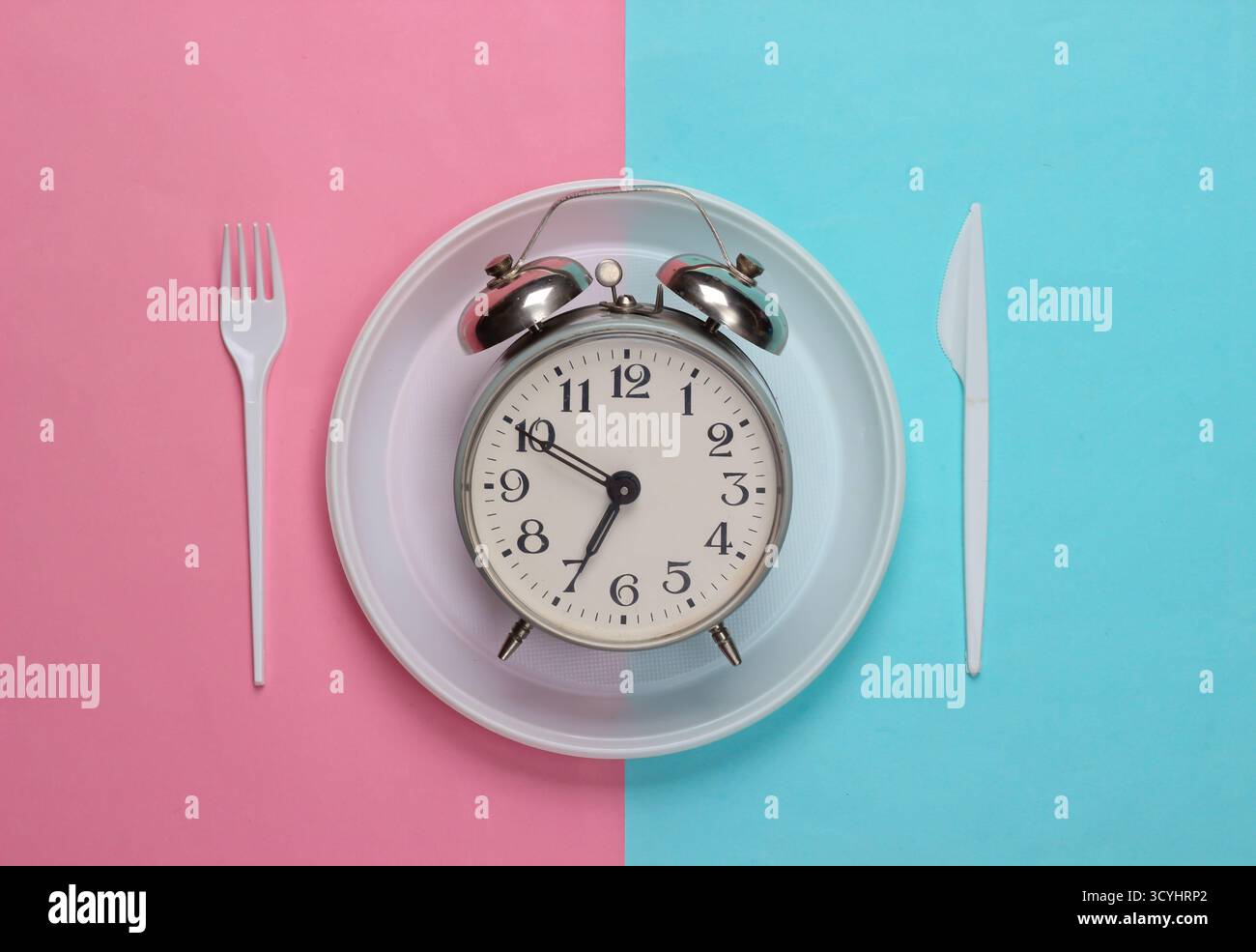 Heure du pique-nique. Réveil dans une assiette avec une fourchette jetable et un couteau sur fond pastel bleu rose. Vue de dessus Banque D'Images