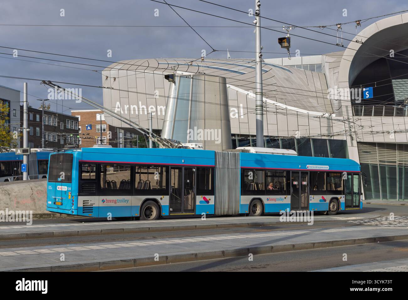 Un long trolleybus articulé bleu passe devant l'architecture moderne et angulaire de la gare centrale d'Arnhem sous le réseau complexe de câbles aériens Banque D'Images
