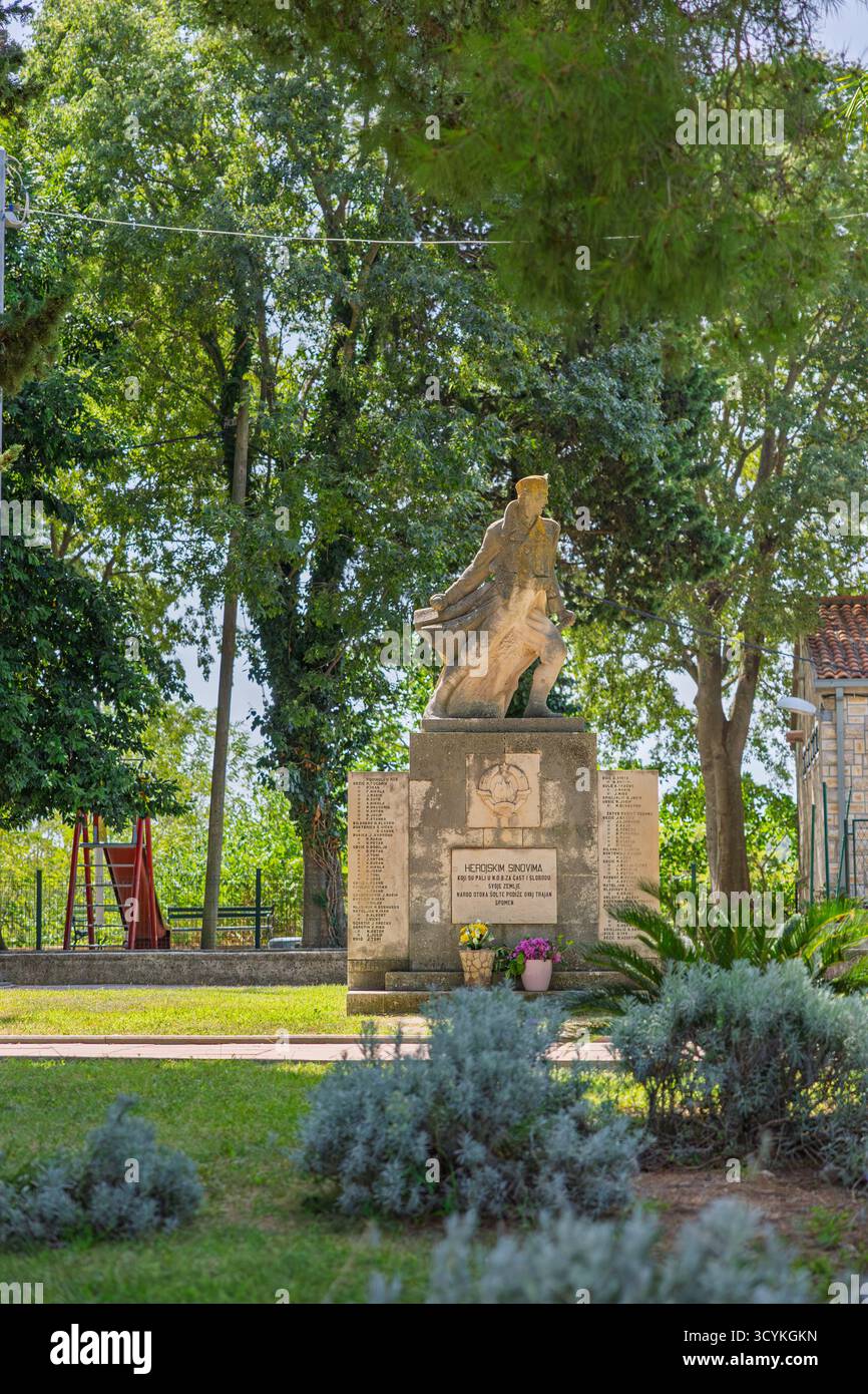 Grohote, CROATIE, 08.29.2025 :Statue en pierre extérieure d'un soldat en marche au sommet d'un piédestal de brique, flanqué de plaques nominatives. Des fleurs fraîches reposent à la base Banque D'Images
