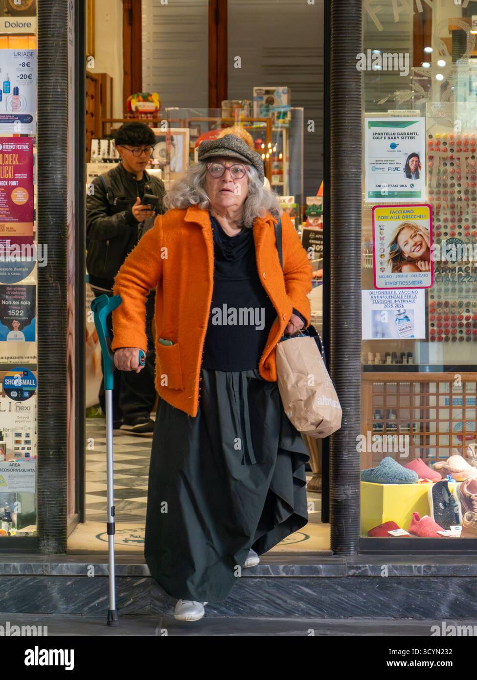 Pise, Italie - 16 octobre 2025 femme âgée avec des cheveux argentés et une veste de corail sortant de la pharmacie Farmacia Bottani dans une rue animée de la ville, tenant un Banque D'Images