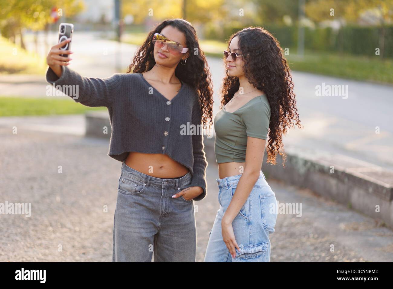 Deux jeunes femmes adultes portant des lunettes de soleil et des vêtements décontractés posent pour un selfie pendant l'heure d'or, capturant un moment d'amitié et de vie moderne Banque D'Images