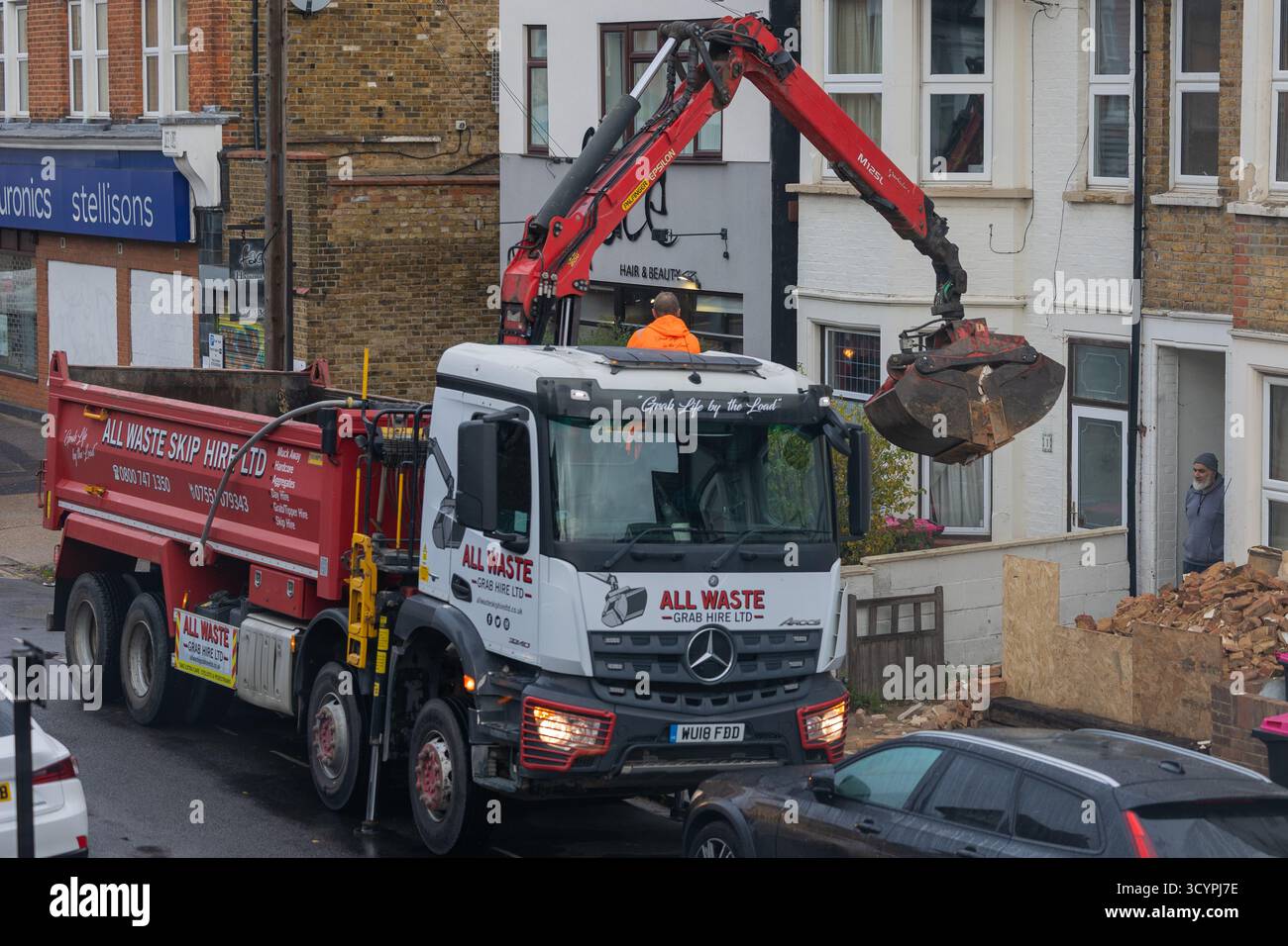 Un camion de location de benne soulève un conteneur métallique rempli de débris via une grue hydraulique, actionnée par un travailleur en orange sur une rue résidentielle bordée de briques. Banque D'Images