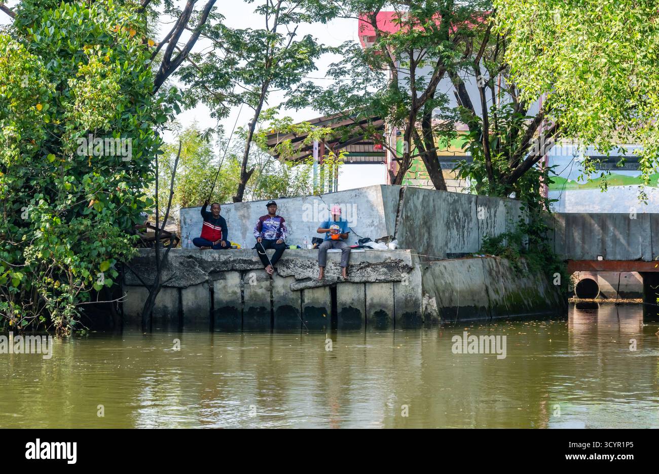Trois hommes pêchant avec des cannes sur un mur de béton près d'une rivière. Java, Indonésie. Banque D'Images