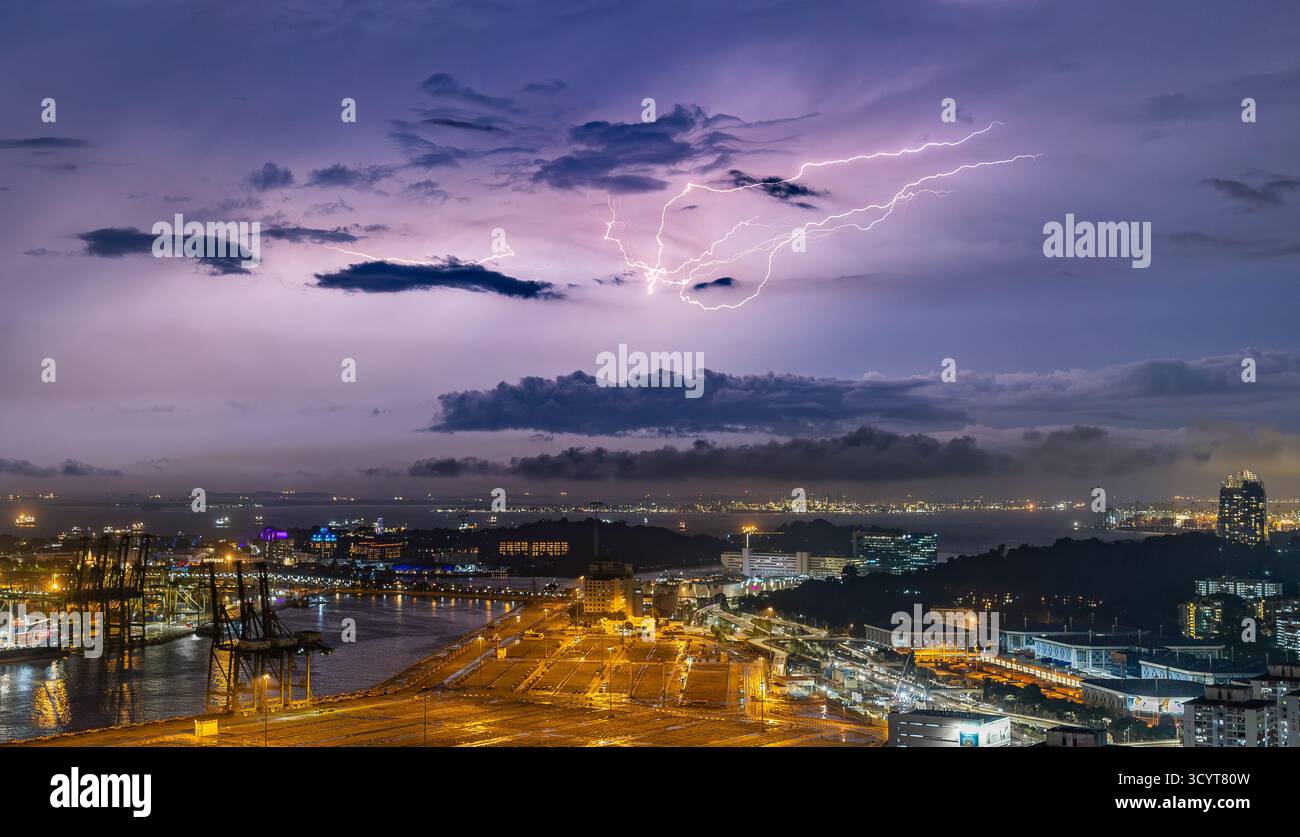 La foudre illumine le ciel nocturne au-dessus de Singapour, avec des grues à conteneurs et des lumières de la ville qui se reflètent sur l'eau en contrebas. Banque D'Images