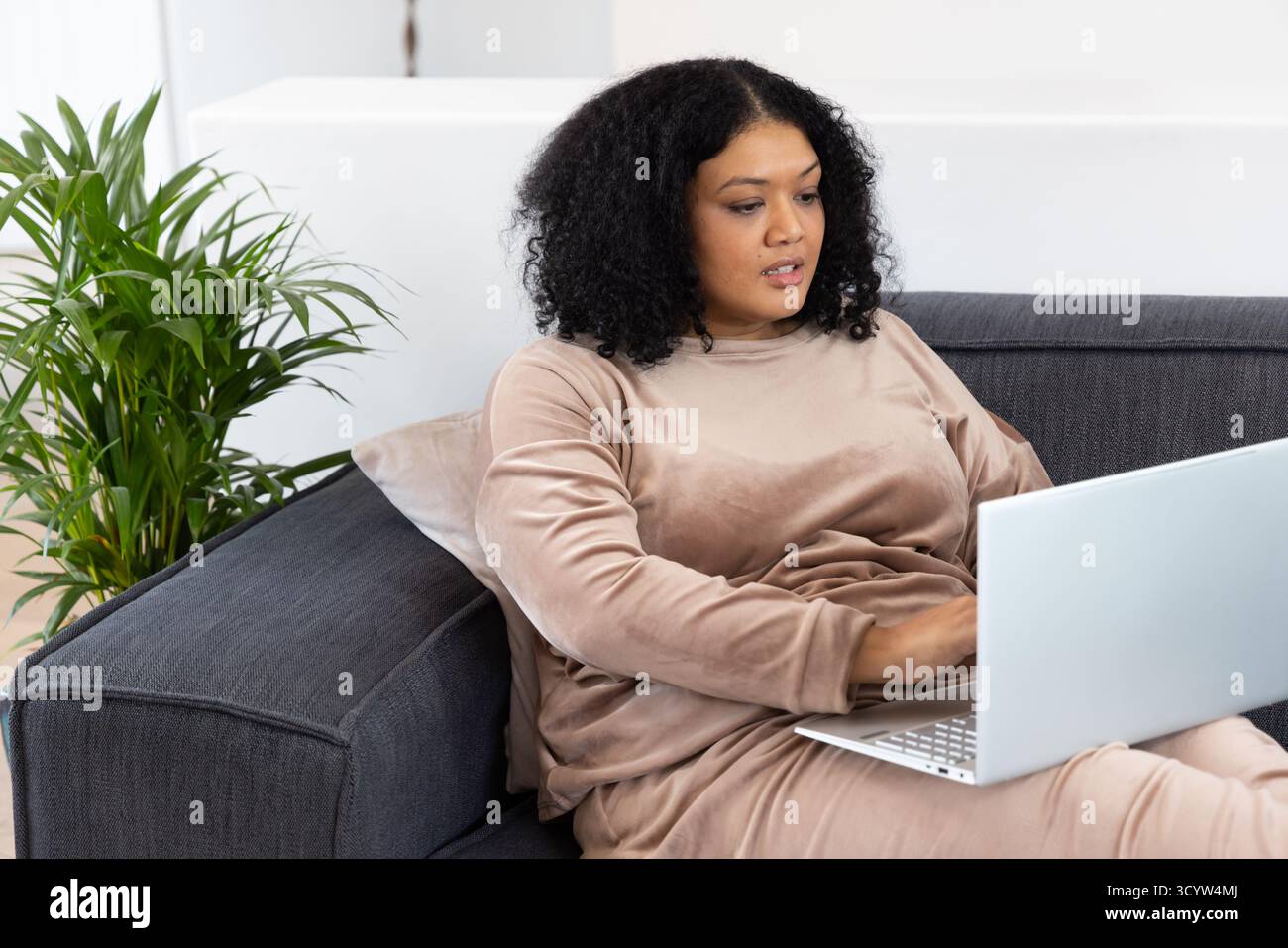 Femme afro-américaine adulte moyenne assise sur le canapé à l'aide d'un ordinateur portable argenté à la maison à côté du coussin beige. Contemporain, minimaliste, cosy, espace de travail, lifestyl Banque D'Images Femme afro-américaine adulte moyenne assise sur le canapé à l'aide d'un ordinateur portable argenté à la maison à côté du coussin beige. Contemporain, minimaliste, cosy, espace de travail, lifestyl Banque D'Images
