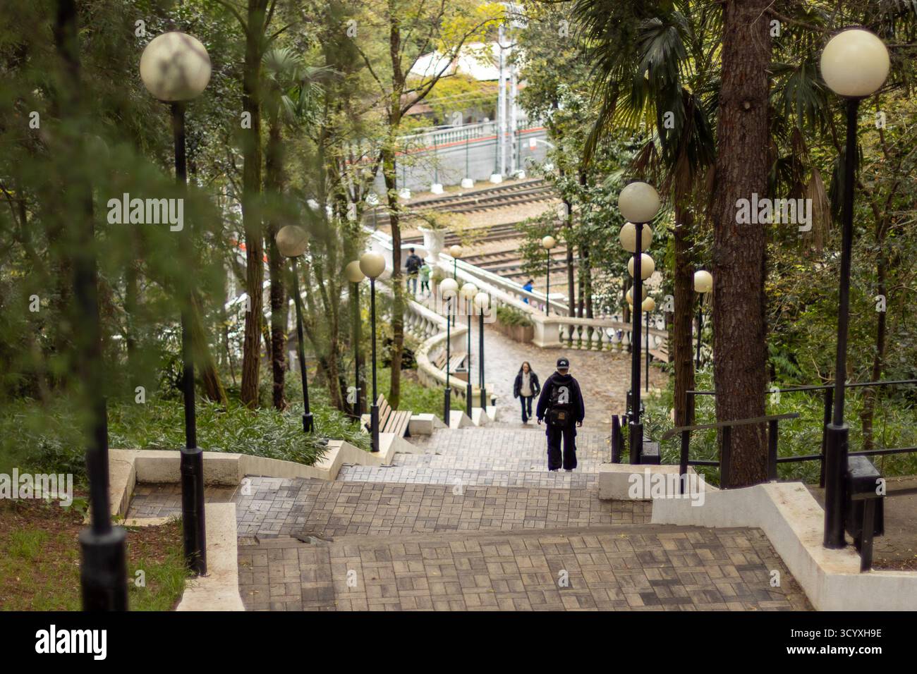 Escaliers pavés dans un parc luxuriant. Escaliers pavés serpentant vers le bas à travers un parc urbain luxuriant avec de grands arbres, des lampes vintage, et des voies ferrées lointaines, ev Banque D'Images