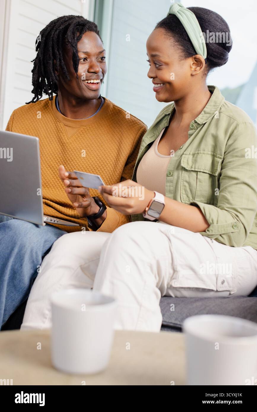 Couple afro-américain assis sur un canapé gris dans le salon à l'aide d'un ordinateur portable et tenant la carte de crédit. Confortable, décontracté, moderne, technologie, domestique, relation Banque D'Images Couple afro-américain assis sur un canapé gris dans le salon à l'aide d'un ordinateur portable et tenant la carte de crédit. Confortable, décontracté, moderne, technologie, domestique, relation Banque D'Images