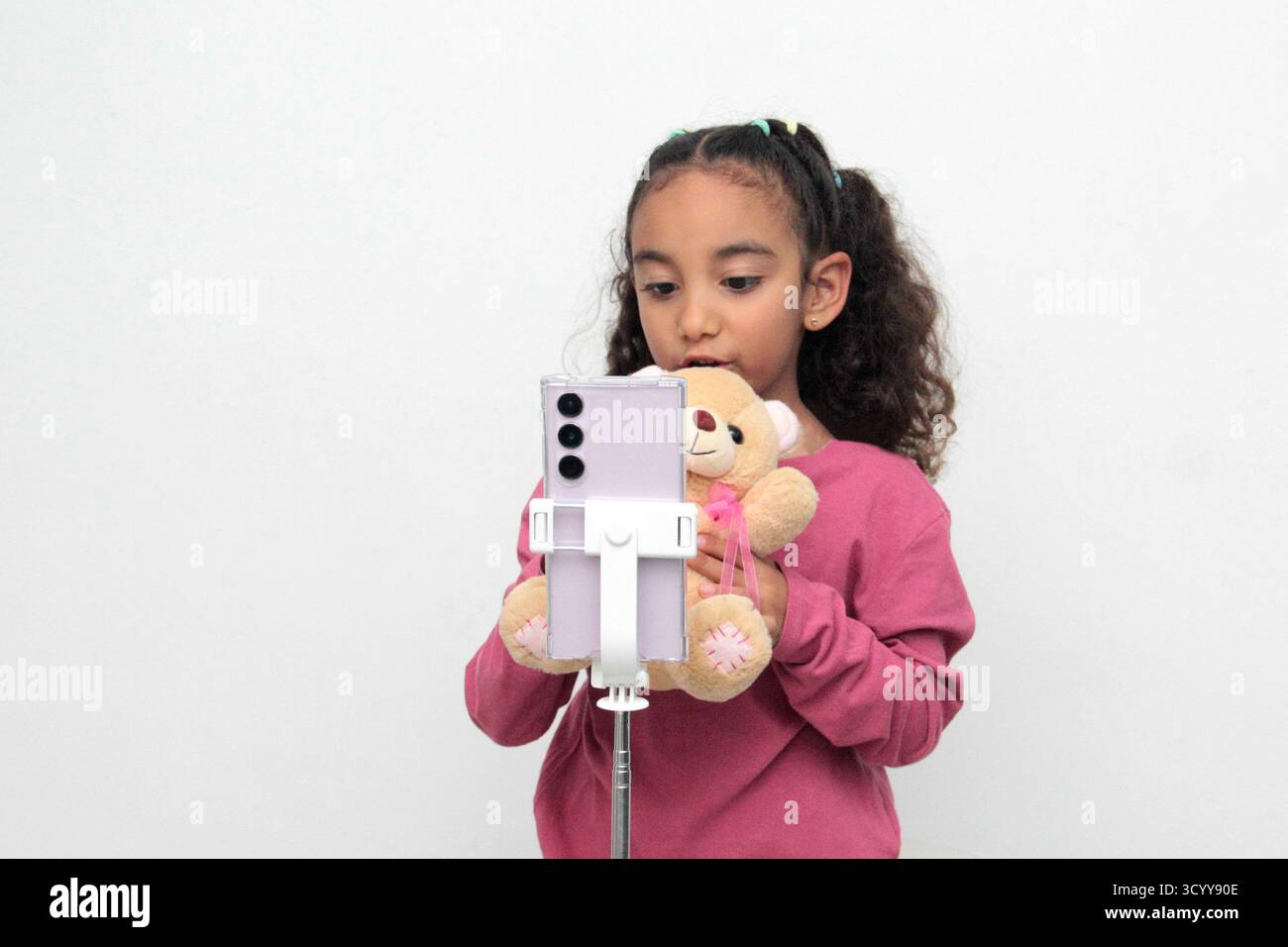 Latina Brunette, 5 ans, enregistre des vidéos avec son téléphone portable sur un trépied, accompagnée de son ours en peluche, pour les publier sur les réseaux sociaux Banque D'Images