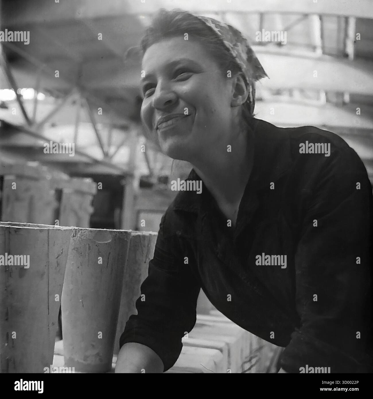 Cette photo d'archive de 1966 de Sloviansk, RSS d'Ukraine, est un portrait réconfortant d'une jeune travailleuse à l'usine d'armature et d'isolateur (AIF). Capturée à son poste de travail, la femme porte une chemise sombre et un foulard à motifs. Elle lève les yeux avec un sourire éclatant et authentique. Devant elle se trouvent plusieurs articles en céramique brute et cylindrique. Le plafond en treillis de l'usine est visible en arrière-plan. Cette image capture l'esprit positif et le côté humain du travail soviétique, un document du Donbass pacifique Banque D'Images