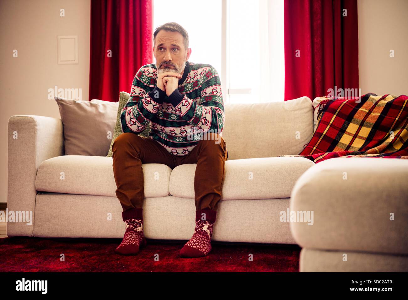 Homme senior se relaxant à la maison pendant la saison de Noël portant un pull festif, montrant une expression réfléchie entourée de décorations chaudes Banque D'Images