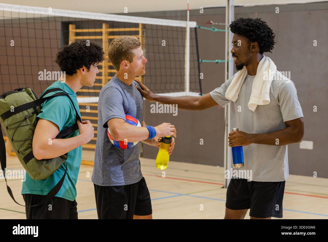 Divers coéquipiers masculins se reposant sur le terrain de volley-ball de gymnastique par filet tenant le volley-ball et des bouteilles d'eau. Athlétique, travail d'équipe, fitness, vêtements de sport, mouvement, camaraderie, énergique || modèle libéré Banque D'Images