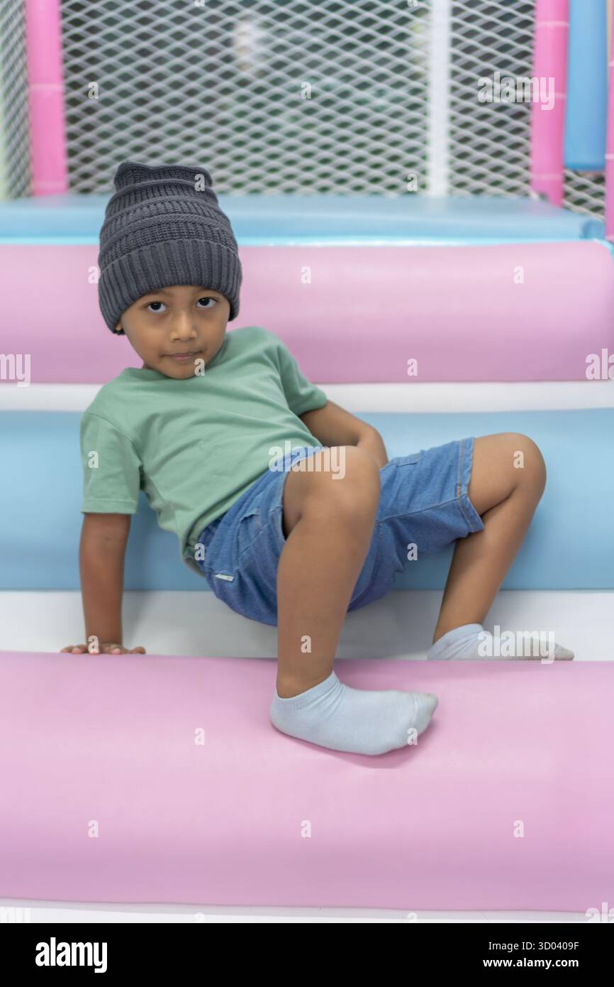 Enfant asiatique heureux posant sur une structure douce lumineuse entourée de coussins pastel vifs, souriant et jouissant d'une activité intérieure ludique. Banque D'Images