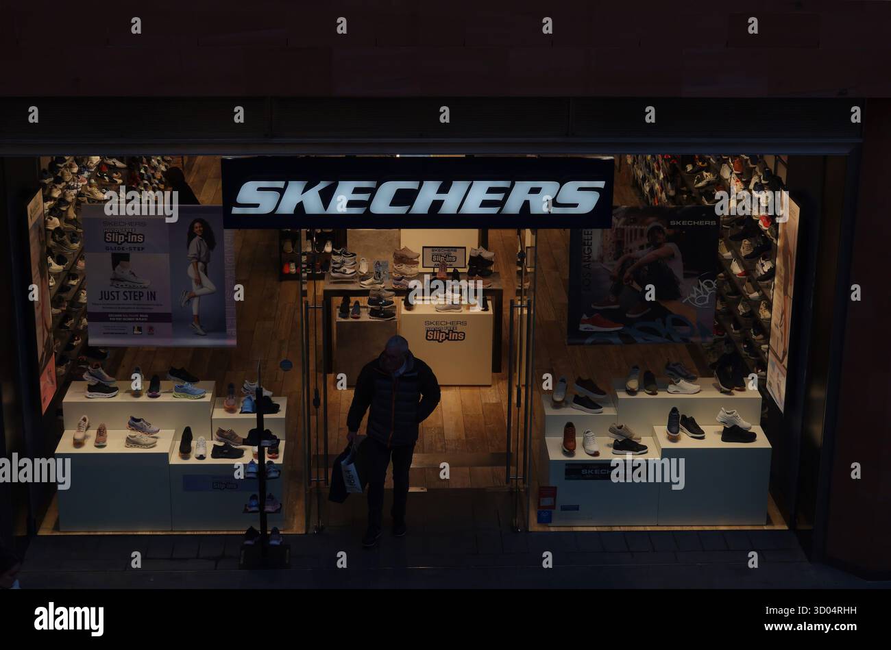 Magasin de chaussures Skechers avec enseignes lumineuses, présentoirs de baskets, et un acheteur sortant tenant un sac à provisions. Banque D'Images