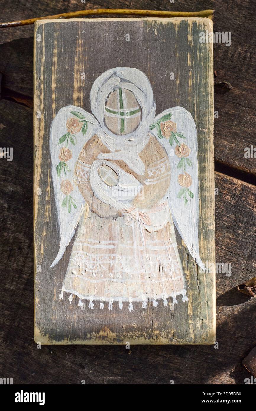 Icône en bois rustique peinte à la main représentant un ange stylisé avec des ailes florales, reposant sur une surface en bois altérée à la lumière naturelle du soleil. Banque D'Images