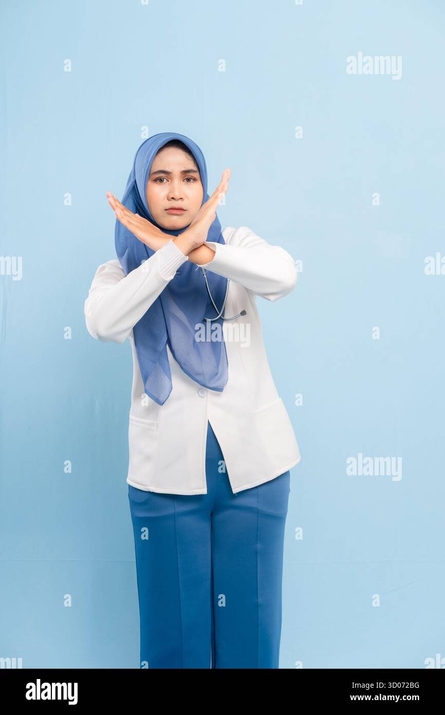 Portrait d'un médecin ou d'une infirmière musulmane asiatique sérieuse montrant un geste d'arrêt ou de non avec les bras croisés, isolé sur un fond bleu. Banque D'Images