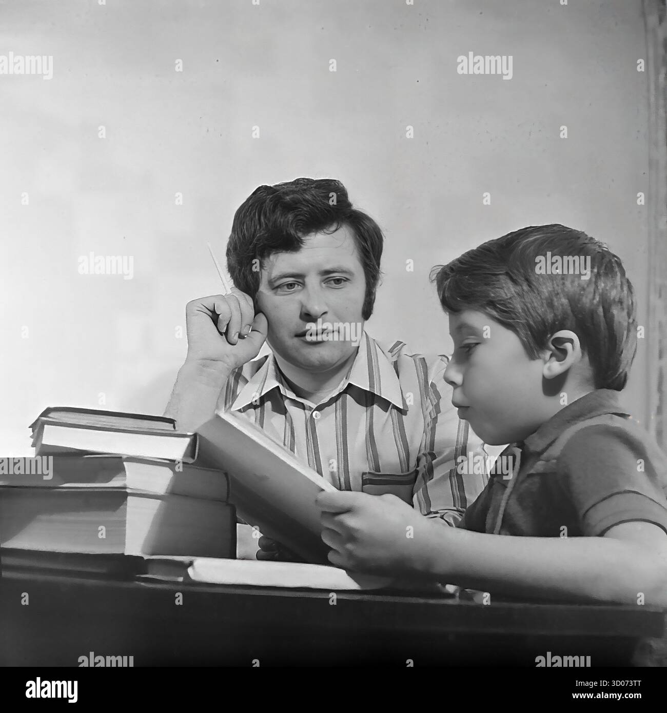 Un père soviétique, un machiniste, regarde attentivement son fils lire un livre chez lui à Sloviansk, dans les années 1970-1980 Cette photo d'archives en noir et blanc de l'URSS, RSS d'Ukraine, est un portrait intime de père et de fils. L'homme, dans une chemise rayée, tient une cigarette tout en observant le garçon, qui est concentré sur ses études. Une grande pile de livres se trouve sur la table polie. Cette image capture une scène de la vie soviétique quotidienne, de la paternité, des valeurs familiales et de l'éducation à la maison. Un document nostalgique de la vie paisible dans le Donbass avant la guerre. Banque D'Images Un père soviétique, un machiniste, regarde attentivement son fils lire un livre chez lui à Sloviansk, dans les années 1970-1980 Cette photo d'archives en noir et blanc de l'URSS, RSS d'Ukraine, est un portrait intime de père et de fils. L'homme, dans une chemise rayée, tient une cigarette tout en observant le garçon, qui est concentré sur ses études. Une grande pile de livres se trouve sur la table polie. Cette image capture une scène de la vie soviétique quotidienne, de la paternité, des valeurs familiales et de l'éducation à la maison. Un document nostalgique de la vie paisible dans le Donbass avant la guerre. Banque D'Images