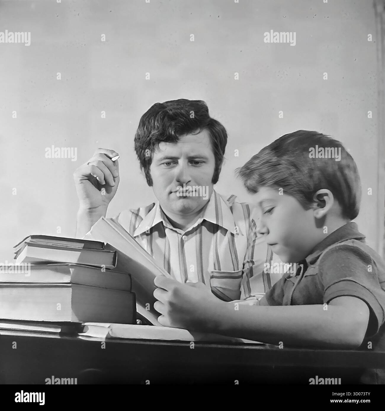 Un père soviétique, un machiniste, regarde attentivement son fils lire un livre chez lui à Sloviansk, dans les années 1970-1980 Cette photo d'archives en noir et blanc de l'URSS, RSS d'Ukraine, est un portrait intime de père et de fils. L'homme, dans une chemise rayée, tient une cigarette tout en observant le garçon, qui est concentré sur ses études. Une grande pile de livres se trouve sur la table polie. Cette image capture une scène de la vie soviétique quotidienne, de la paternité, des valeurs familiales et de l'éducation à la maison. Un document nostalgique de la vie paisible dans le Donbass avant la guerre. Banque D'Images Un père soviétique, un machiniste, regarde attentivement son fils lire un livre chez lui à Sloviansk, dans les années 1970-1980 Cette photo d'archives en noir et blanc de l'URSS, RSS d'Ukraine, est un portrait intime de père et de fils. L'homme, dans une chemise rayée, tient une cigarette tout en observant le garçon, qui est concentré sur ses études. Une grande pile de livres se trouve sur la table polie. Cette image capture une scène de la vie soviétique quotidienne, de la paternité, des valeurs familiales et de l'éducation à la maison. Un document nostalgique de la vie paisible dans le Donbass avant la guerre. Banque D'Images