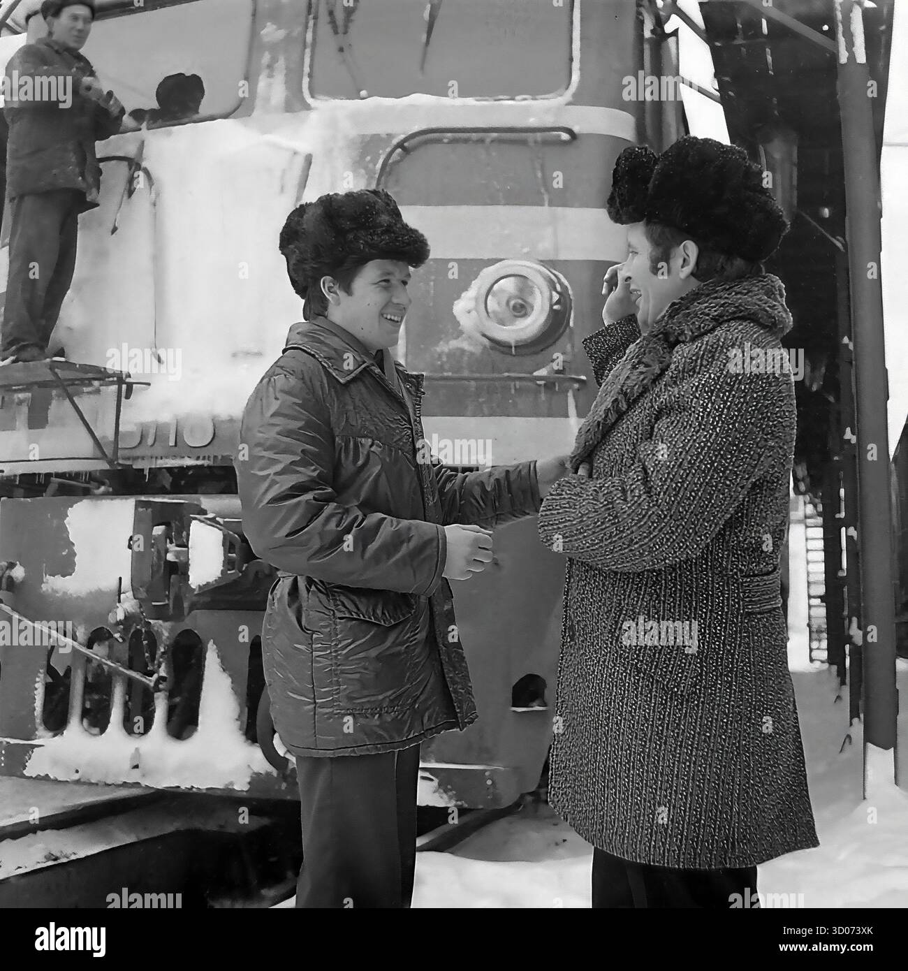Un machiniste soviétique et vétéran (à droite) discute avec un collègue de chemin de fer plus jeune à Sloviansk, dans les années 1970-1980 Archive photo en noir et blanc, URSS, RSS d'Ukraine. Ils se trouvent devant une grande locomotive diesel (peut-être ChME3) dans un dépôt ferroviaire. Les deux hommes portent des manteaux d'hiver et des chapeaux de fourrure ('papakha'). En arrière-plan, un autre travailleur nettoie le train, qui est couvert de neige et de glaçons. Cette image montre la vie industrielle soviétique quotidienne, la camaraderie des ouvriers et un hiver rigoureux. Un document précieux de la vie paisible dans le Donbass avant la guerre. Banque D'Images Un machiniste soviétique et vétéran (à droite) discute avec un collègue de chemin de fer plus jeune à Sloviansk, dans les années 1970-1980 Archive photo en noir et blanc, URSS, RSS d'Ukraine. Ils se trouvent devant une grande locomotive diesel (peut-être ChME3) dans un dépôt ferroviaire. Les deux hommes portent des manteaux d'hiver et des chapeaux de fourrure ('papakha'). En arrière-plan, un autre travailleur nettoie le train, qui est couvert de neige et de glaçons. Cette image montre la vie industrielle soviétique quotidienne, la camaraderie des ouvriers et un hiver rigoureux. Un document précieux de la vie paisible dans le Donbass avant la guerre. Banque D'Images