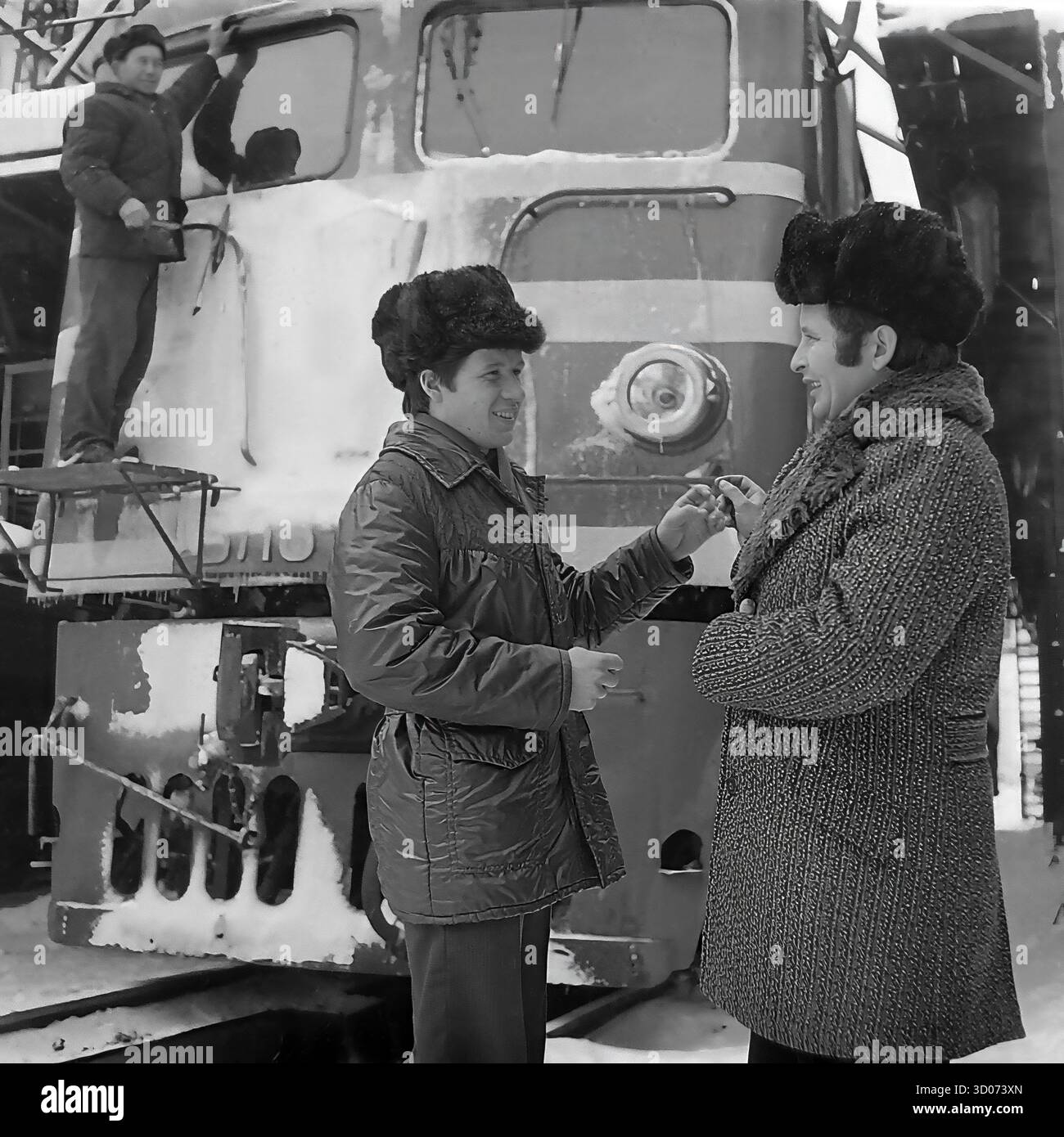 Un machiniste soviétique et vétéran (à droite) discute avec un collègue de chemin de fer plus jeune à Sloviansk, dans les années 1970-1980 Archive photo en noir et blanc, URSS, RSS d'Ukraine. Ils se trouvent devant une grande locomotive diesel (peut-être ChME3) dans un dépôt ferroviaire. Les deux hommes portent des manteaux d'hiver et des chapeaux de fourrure ('papakha'). En arrière-plan, un autre travailleur nettoie le train, qui est couvert de neige et de glaçons. Cette image montre la vie industrielle soviétique quotidienne, la camaraderie des ouvriers et un hiver rigoureux. Un document précieux de la vie paisible dans le Donbass avant la guerre. Banque D'Images Un machiniste soviétique et vétéran (à droite) discute avec un collègue de chemin de fer plus jeune à Sloviansk, dans les années 1970-1980 Archive photo en noir et blanc, URSS, RSS d'Ukraine. Ils se trouvent devant une grande locomotive diesel (peut-être ChME3) dans un dépôt ferroviaire. Les deux hommes portent des manteaux d'hiver et des chapeaux de fourrure ('papakha'). En arrière-plan, un autre travailleur nettoie le train, qui est couvert de neige et de glaçons. Cette image montre la vie industrielle soviétique quotidienne, la camaraderie des ouvriers et un hiver rigoureux. Un document précieux de la vie paisible dans le Donbass avant la guerre. Banque D'Images