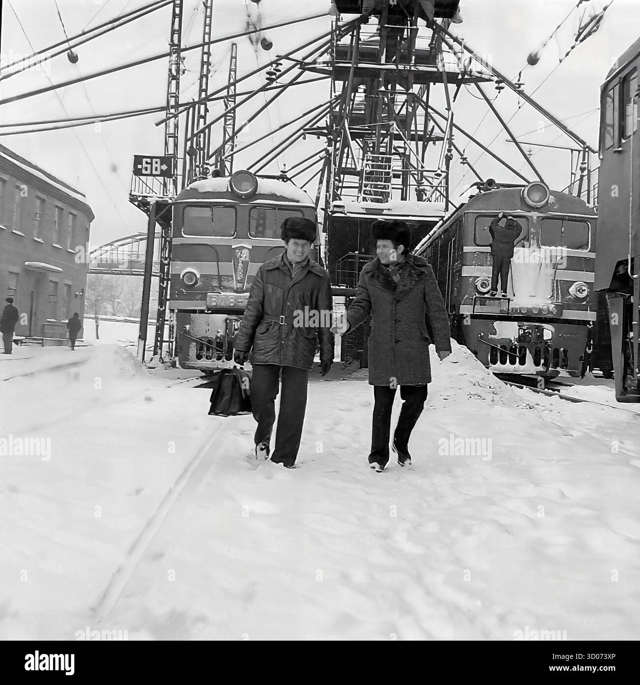 Deux cheminots soviétiques, dont un machiniste et vétéran (à droite), traversent un dépôt de chemin de fer enneigé à Sloviansk, dans les années 1970-1980 Archive photo en noir et blanc, URSS, RSS d'Ukraine. Les hommes, portant des manteaux d'hiver et des chapeaux de fourrure, marchent et parlent, avec un portant une mallette. En arrière-plan, on trouve des locomotives électriques soviétiques, dont un VL8 (ВЛ8-701), et une infrastructure ferroviaire complexe. Cette image montre la vie industrielle soviétique quotidienne, la profession de transport et les travailleurs dans des conditions hivernales difficiles. Un document précieux de la vie paisible dans le Donbass avant la guerre. Banque D'Images Deux cheminots soviétiques, dont un machiniste et vétéran (à droite), traversent un dépôt de chemin de fer enneigé à Sloviansk, dans les années 1970-1980 Archive photo en noir et blanc, URSS, RSS d'Ukraine. Les hommes, portant des manteaux d'hiver et des chapeaux de fourrure, marchent et parlent, avec un portant une mallette. En arrière-plan, on trouve des locomotives électriques soviétiques, dont un VL8 (ВЛ8-701), et une infrastructure ferroviaire complexe. Cette image montre la vie industrielle soviétique quotidienne, la profession de transport et les travailleurs dans des conditions hivernales difficiles. Un document précieux de la vie paisible dans le Donbass avant la guerre. Banque D'Images