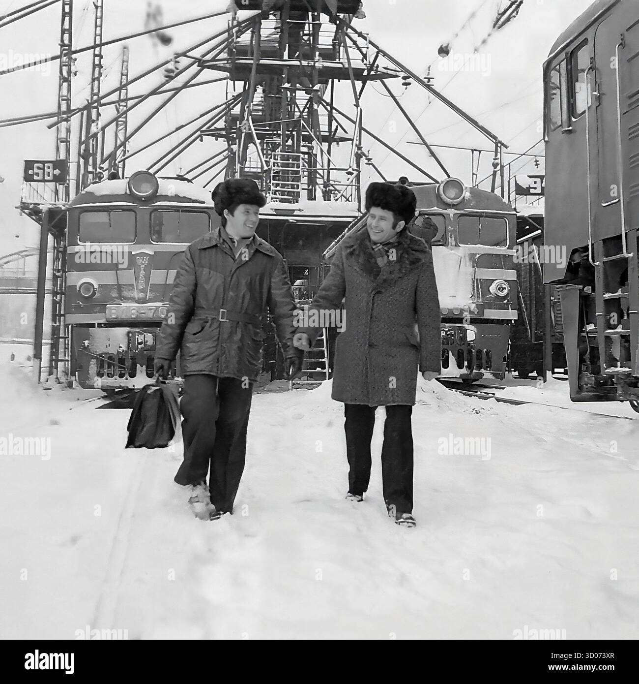 Deux cheminots soviétiques, dont un machiniste et vétéran (à droite), traversent un dépôt de chemin de fer enneigé à Sloviansk, dans les années 1970-1980 Archive photo en noir et blanc, URSS, RSS d'Ukraine. Les hommes, portant des manteaux d'hiver et des chapeaux de fourrure, marchent et parlent, avec un portant une mallette. En arrière-plan, on trouve des locomotives électriques soviétiques, dont un VL8 (ВЛ8-701), et une infrastructure ferroviaire complexe. Cette image montre la vie industrielle soviétique quotidienne, la profession de transport et les travailleurs dans des conditions hivernales difficiles. Un document précieux de la vie paisible dans le Donbass avant la guerre. Banque D'Images Deux cheminots soviétiques, dont un machiniste et vétéran (à droite), traversent un dépôt de chemin de fer enneigé à Sloviansk, dans les années 1970-1980 Archive photo en noir et blanc, URSS, RSS d'Ukraine. Les hommes, portant des manteaux d'hiver et des chapeaux de fourrure, marchent et parlent, avec un portant une mallette. En arrière-plan, on trouve des locomotives électriques soviétiques, dont un VL8 (ВЛ8-701), et une infrastructure ferroviaire complexe. Cette image montre la vie industrielle soviétique quotidienne, la profession de transport et les travailleurs dans des conditions hivernales difficiles. Un document précieux de la vie paisible dans le Donbass avant la guerre. Banque D'Images