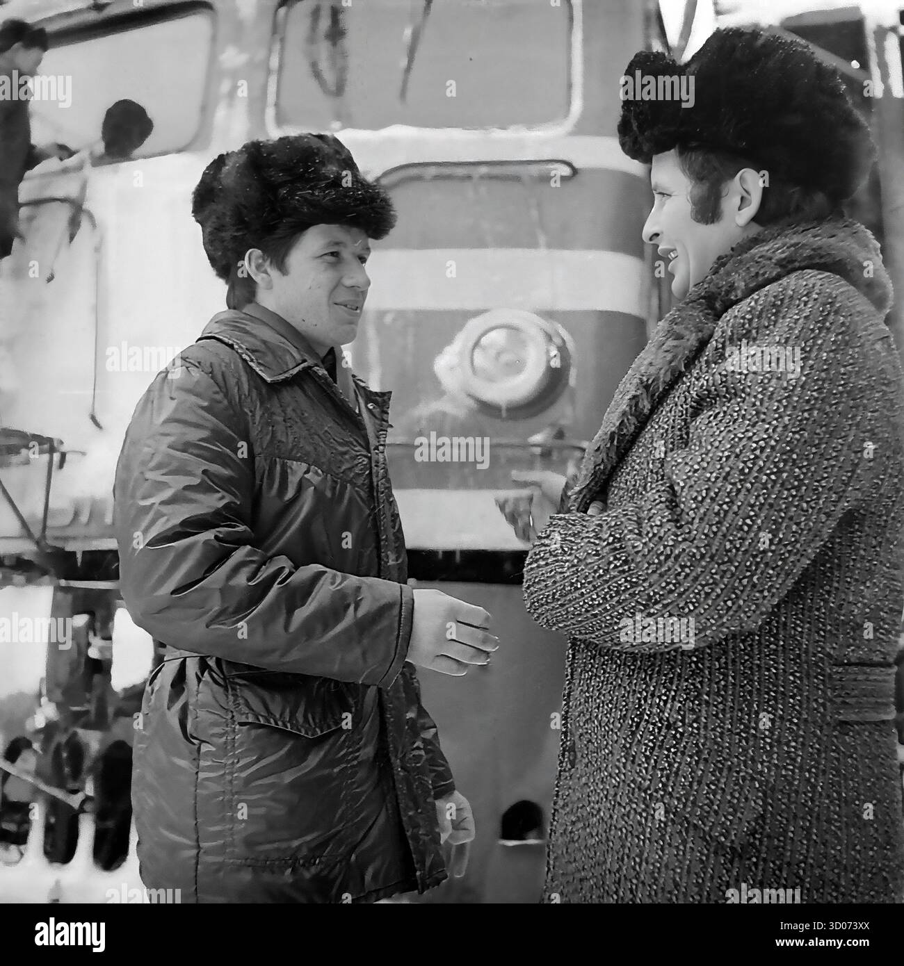Un machiniste soviétique et vétéran (à droite) discute avec un collègue de chemin de fer plus jeune à Sloviansk, dans les années 1970-1980 Archive photo en noir et blanc, URSS, RSS d'Ukraine. Ils se trouvent devant une grande locomotive diesel (peut-être ChME3) dans un dépôt ferroviaire. Les deux hommes portent des manteaux d'hiver et des chapeaux de fourrure ('papakha'). En arrière-plan, un autre travailleur nettoie le train, qui est couvert de neige et de glaçons. Cette image montre la vie industrielle soviétique quotidienne, la camaraderie des ouvriers et un hiver rigoureux. Un document précieux de la vie paisible dans le Donbass avant la guerre. Banque D'Images Un machiniste soviétique et vétéran (à droite) discute avec un collègue de chemin de fer plus jeune à Sloviansk, dans les années 1970-1980 Archive photo en noir et blanc, URSS, RSS d'Ukraine. Ils se trouvent devant une grande locomotive diesel (peut-être ChME3) dans un dépôt ferroviaire. Les deux hommes portent des manteaux d'hiver et des chapeaux de fourrure ('papakha'). En arrière-plan, un autre travailleur nettoie le train, qui est couvert de neige et de glaçons. Cette image montre la vie industrielle soviétique quotidienne, la camaraderie des ouvriers et un hiver rigoureux. Un document précieux de la vie paisible dans le Donbass avant la guerre. Banque D'Images
